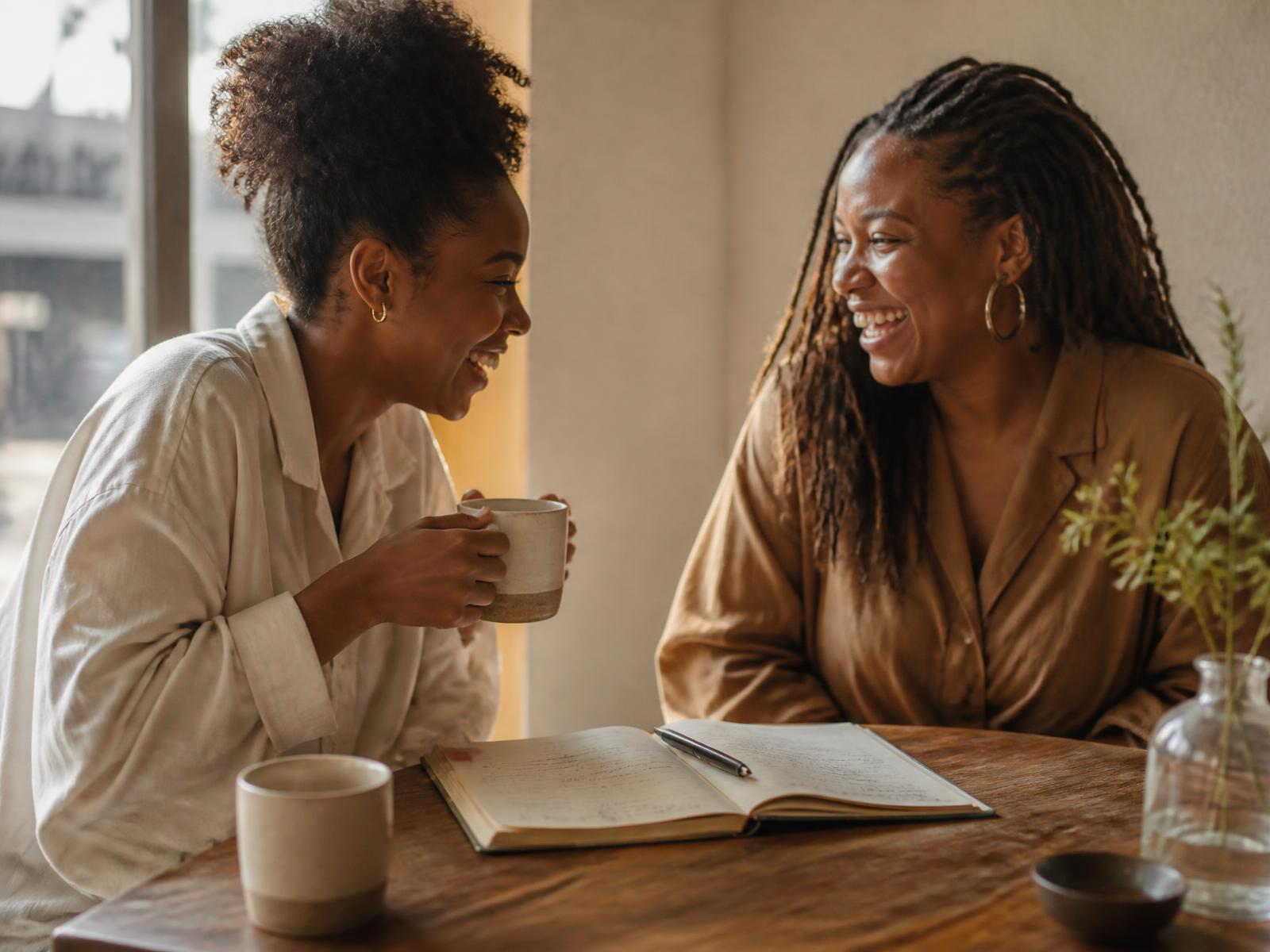 Two Black women laughing together over coffee and an open notebook at a sunlit table